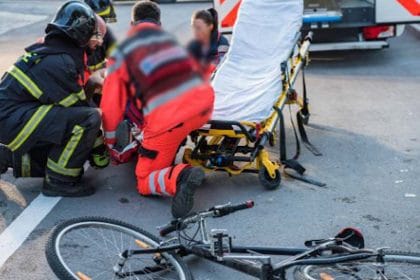 Emergency responders kneel beside a patient on a stretcher on a roadway, with a bicycle lying on the pavement nearby.