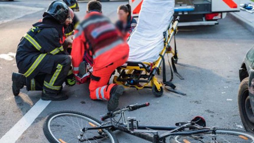 Emergency responders kneel beside a patient on a stretcher on a roadway, with a bicycle lying on the pavement nearby.