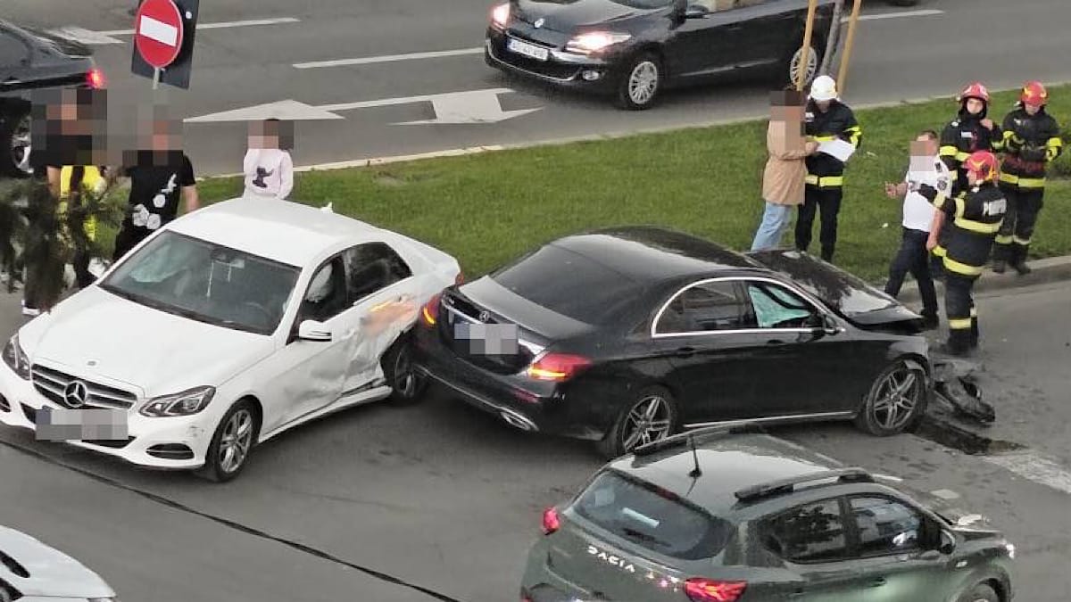 White car and black sedan collide at an intersection; firefighters and bystanders assess the scene. Accident pitesti