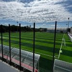 Wide view of an outdoor soccer field with artificial turf, fenced perimeter, and empty stands along the right under a cloudy sky baza sportiva pitesti anghel saligny