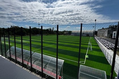 Wide view of an outdoor soccer field with artificial turf, fenced perimeter, and empty stands along the right under a cloudy sky baza sportiva pitesti anghel saligny