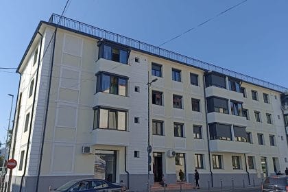 Modern four-story apartment building with balconies along a tree-lined street under a clear blue sky, with parked cars nearby