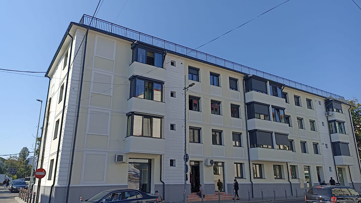 Bloc Pitesti Stiripitesti.ro Modern four-story apartment building with balconies along a tree-lined street under a clear blue sky, with parked cars nearby bloc pitesti